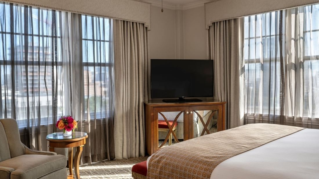 Flat-screen TV on a wooden stand by a chair under drapes in Corner King at Warwick Melrose Dallas