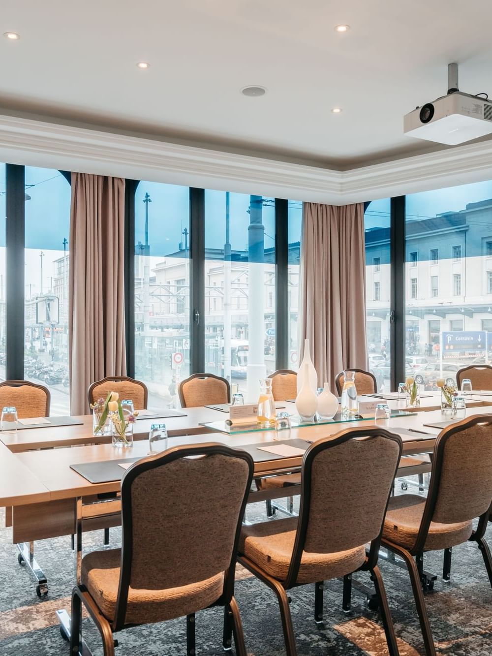Long wooden table by chairs under a ceiling projector near large city windows in Rigi at Warwick Geneva