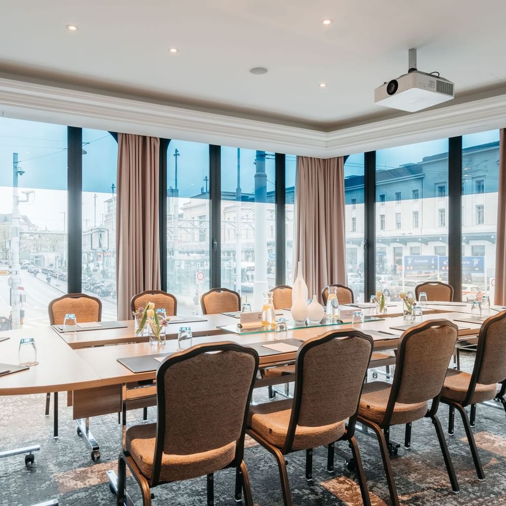 Long wooden table by chairs under a ceiling projector near large city windows in Rigi at Warwick Geneva