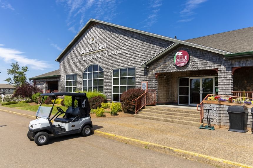 Motorized golf cart parked by a shingled clubhouse entrance under a clear blue sky at Rodd Brudenell River Resort