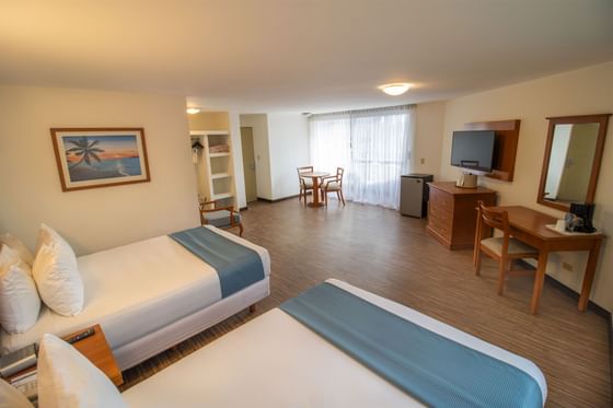 Suite luminosa con dos camas, escritorio y mesa de comedor junto a grandes ventanas en Junior Suite de Araiza Palmira