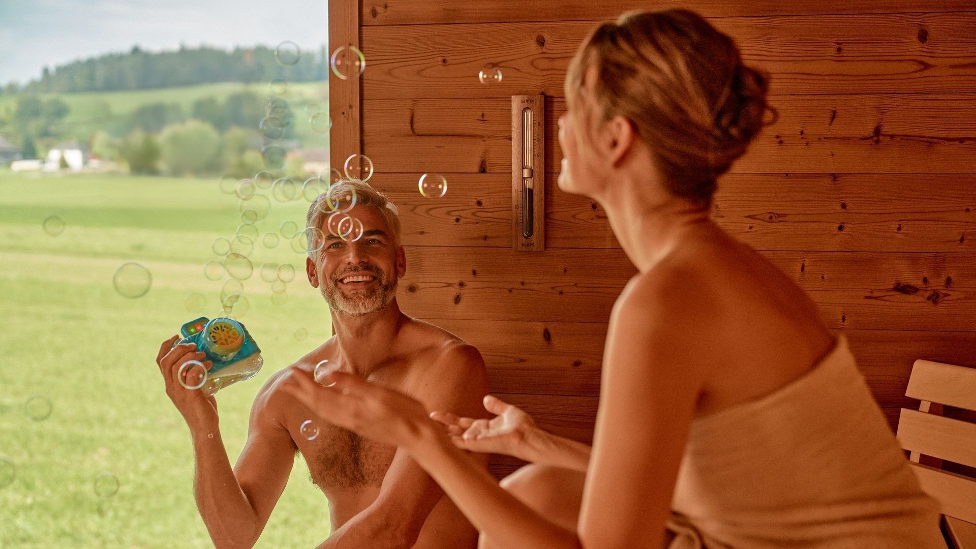 Uomo e donna in sauna con bolle di sapone e vista su un prato verde.