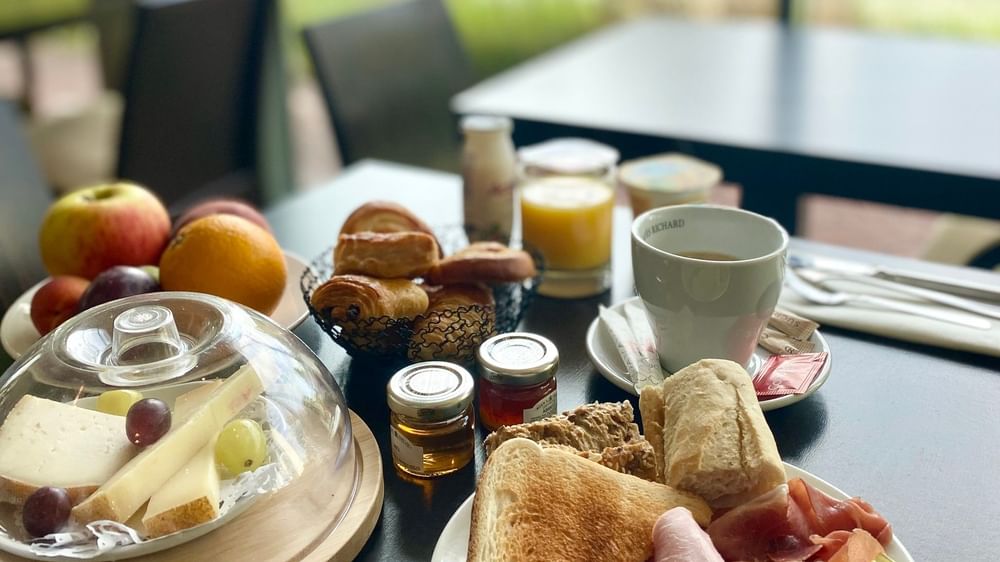 Plateau de petit-déjeuner avec fruits, pain, charcuterie, fromage et boissons chaudes sur une table.