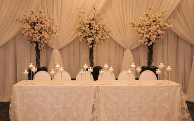 Candles on long table covered in lace cloth before white drapes and pink floral trees at Spencer Hotel & Conference Centre