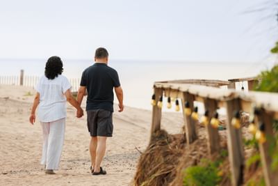 Couple from behind walking hand-in-hand on sandy beach path with wood railing at Rodd Crowbush Golf & Beach Resort
