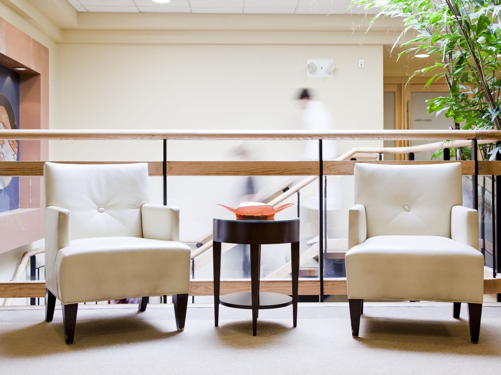 Two white chairs and a small table with an orange bowl on a balcony with a blurry person walking.