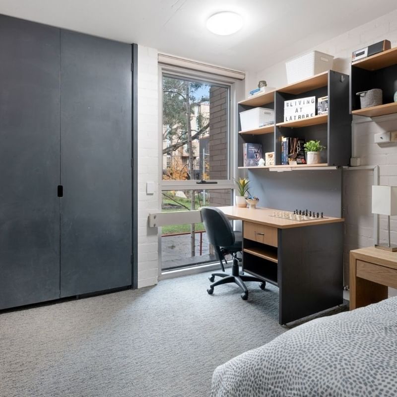 A clean student room with a bed, desk, chair, and shelves at La Trobe University – Menzies College.
