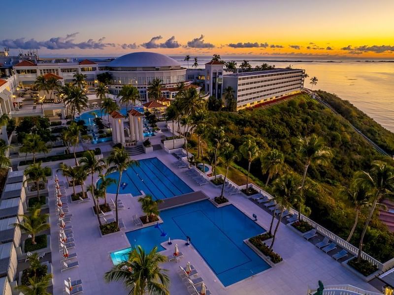 Aerial view of El Conquistador Resort with pools, palm trees, & sunset over the ocean, creating a serene tropical atmosphere