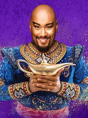 Smiling man dressed in a gold and blue genie costume holds a magic lamp against a purple background at The Sebel Brisbane