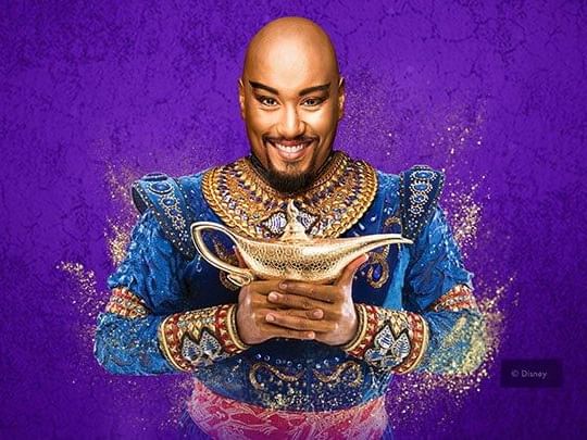 Smiling man dressed in a gold and blue genie costume holds a magic lamp against a purple background at The Sebel Brisbane