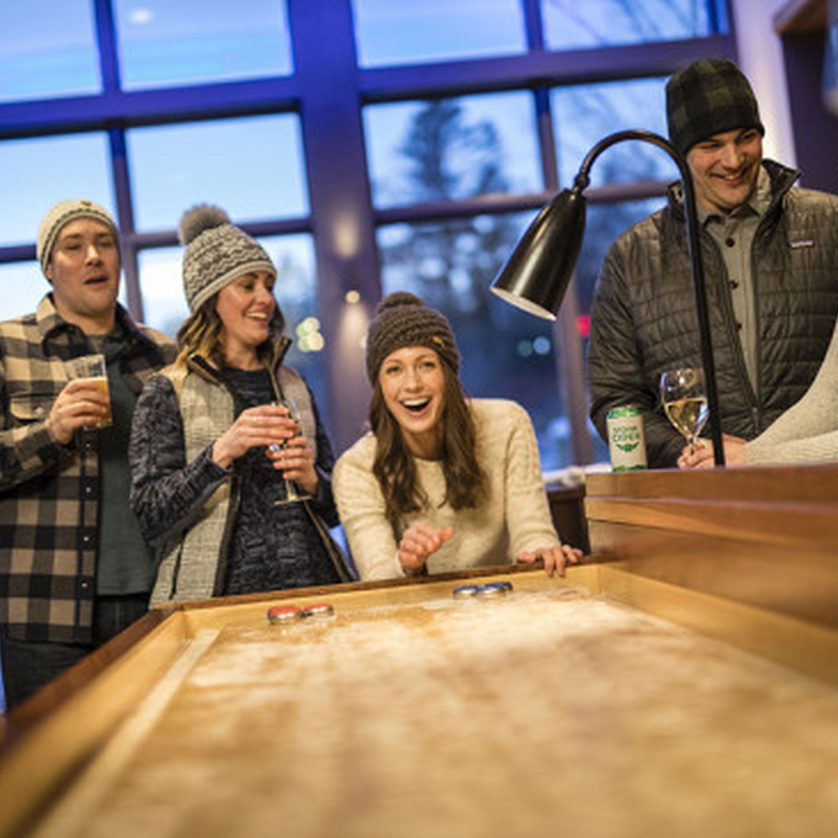 Four people in winter clothes play a game of shuffleboard indoors.