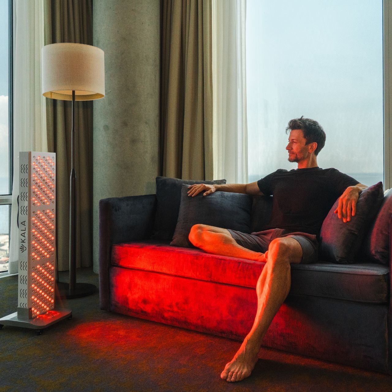 Man sitting on a sofa lit by a glowing red light therapy panel, beside a floor lamp at Hotel X Toronto