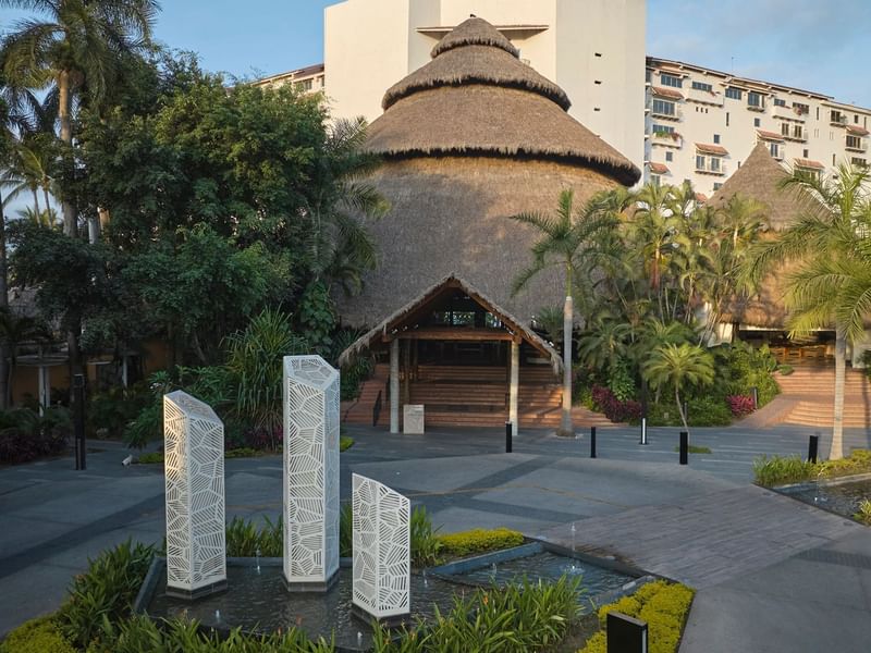 Lush tropical entrance at Fiesta Americana, featuring a large thatched-roof building, palm trees, and modern sculptures