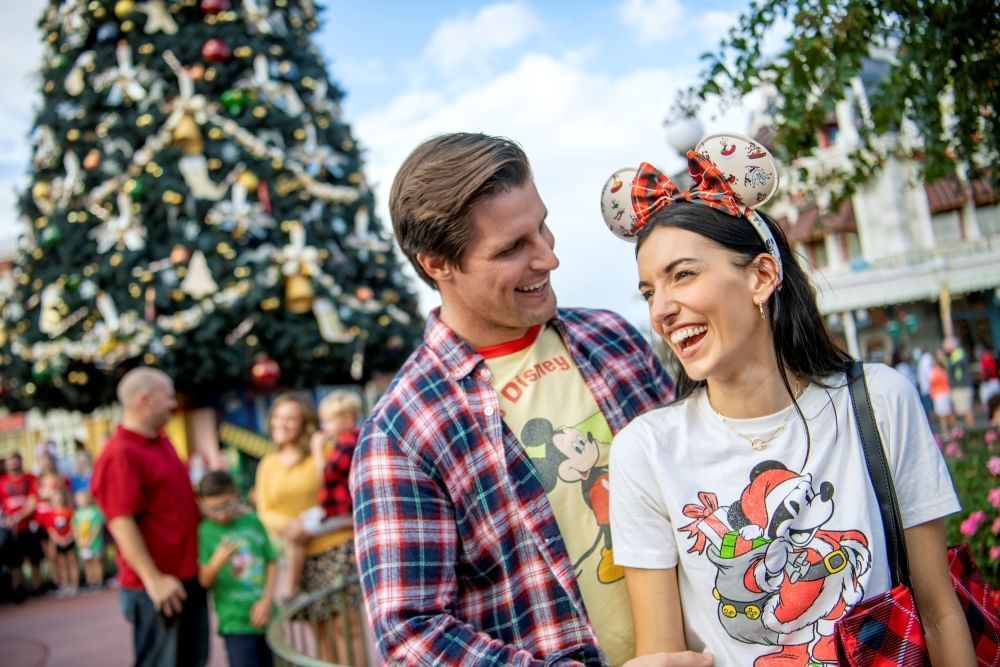 A couple wearing Disney gear laugh at one another in front of a large decorated Christmas tree. 