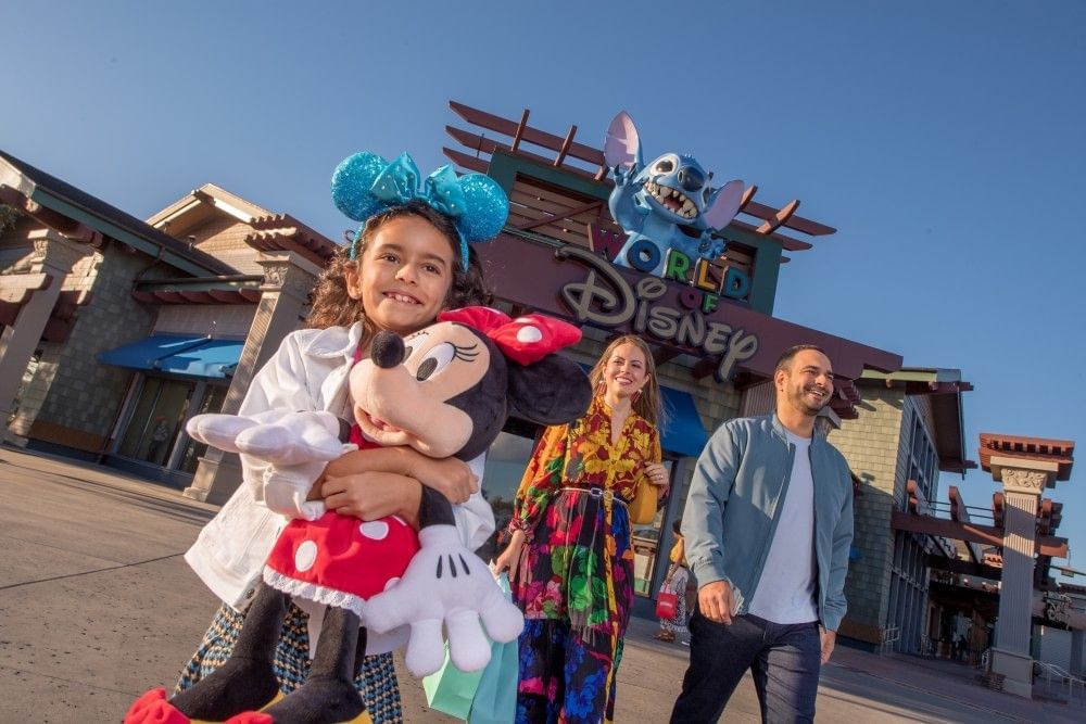 A girl hugs a Minnie Mouse doll as she and her parents leave a large building with a sign that reads World of Disney.