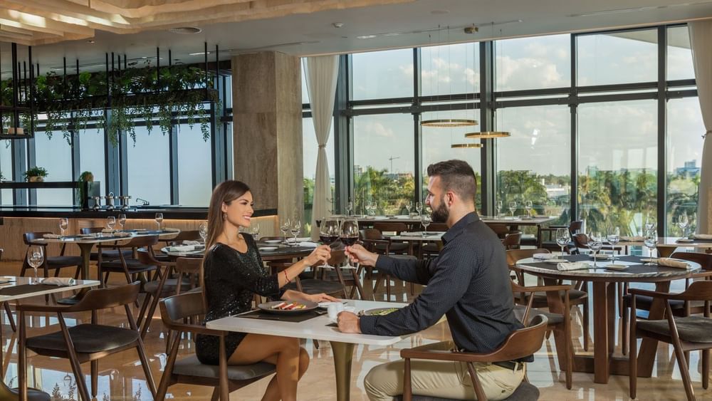 Couple dining in La Huerta restaurant, featuring floor-to-ceiling windows and a modern, airy interior at Camino Real Merida