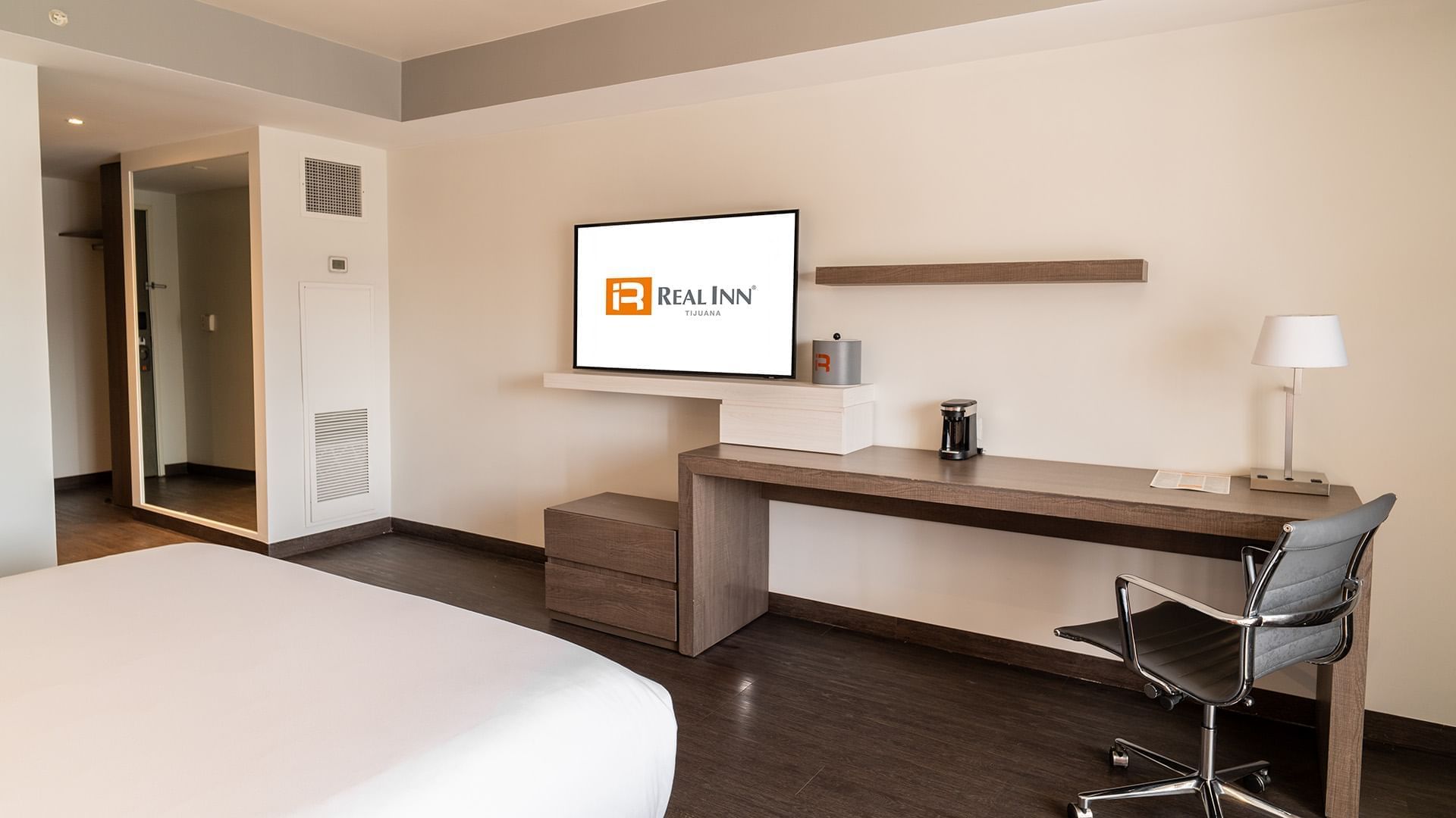 Wall-mounted TV and work desk facing the cozy bed in the King Deluxe Room with wooden floors at Real Inn Tijuana
