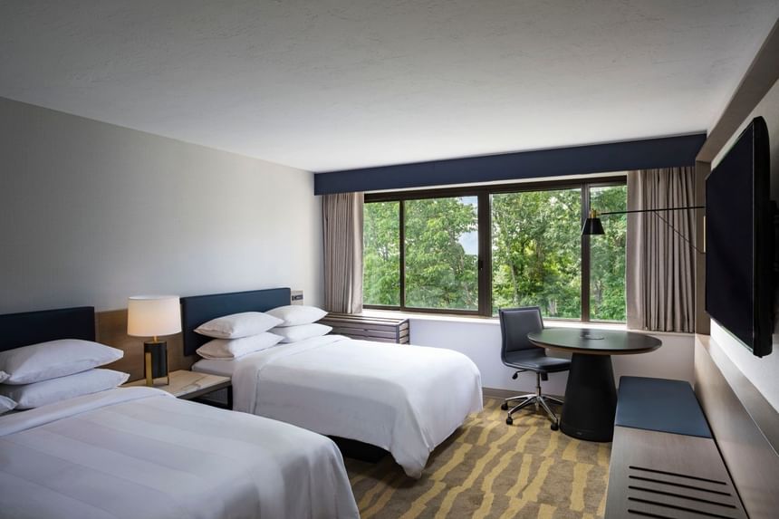 TV facing two beds near a window overlooking the forest in the Double Guest Room at Sable Oaks Weddings and Events