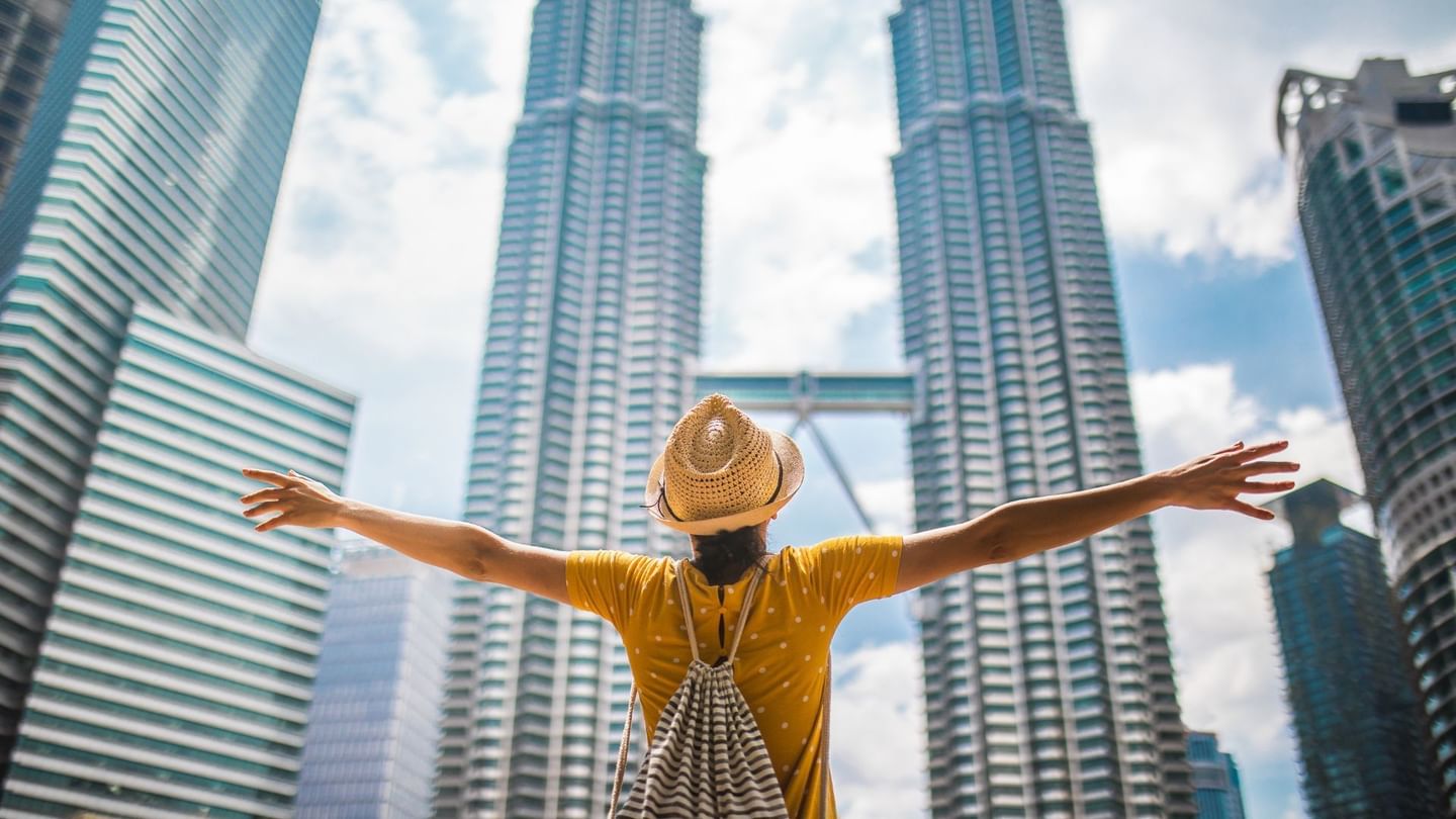 Woman with arms outstretched and hat in front of Twin Petronas Towers and other buildings near Sunway Velocity Hotel