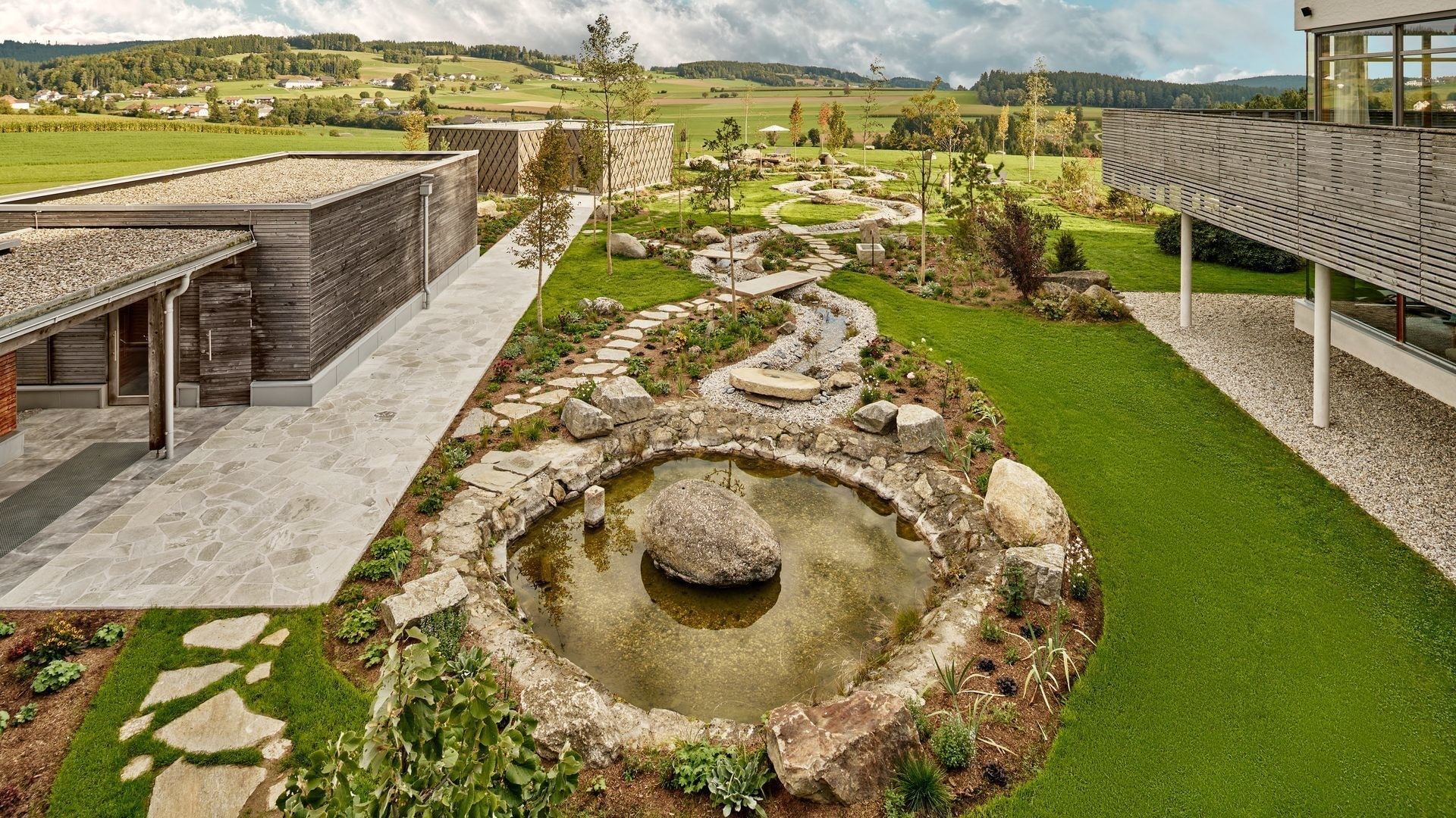 Naturnaher Wellnessgarten mit Teich, Steinwegen und Holzarchitektur vor grüner Hügellandschaft im Falkensteiner Hotel Bad Leonfelden