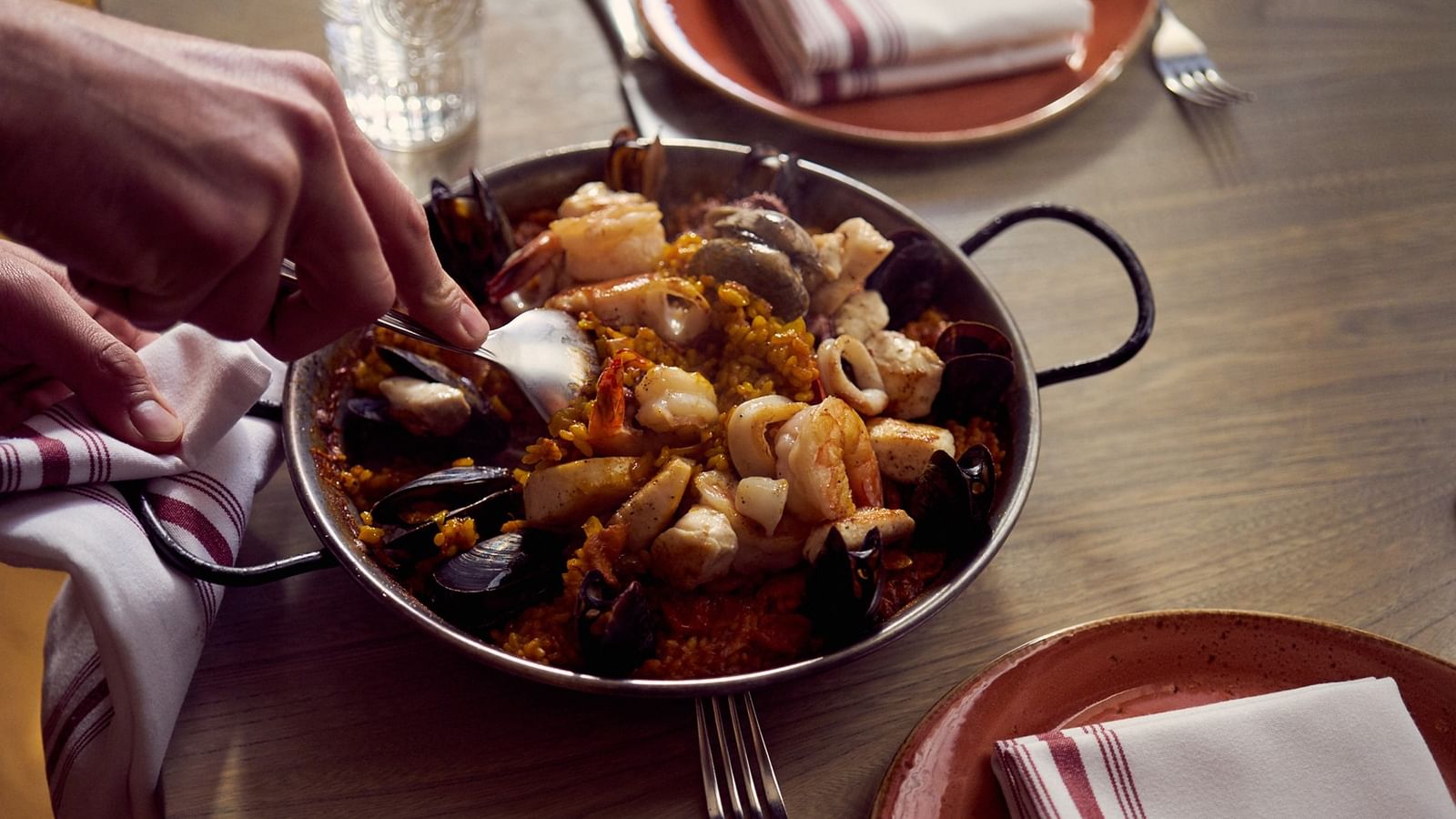 Spoon serving seafood paella in a metal pan by plates with striped napkins on a table at Warwick Hotels & Resorts