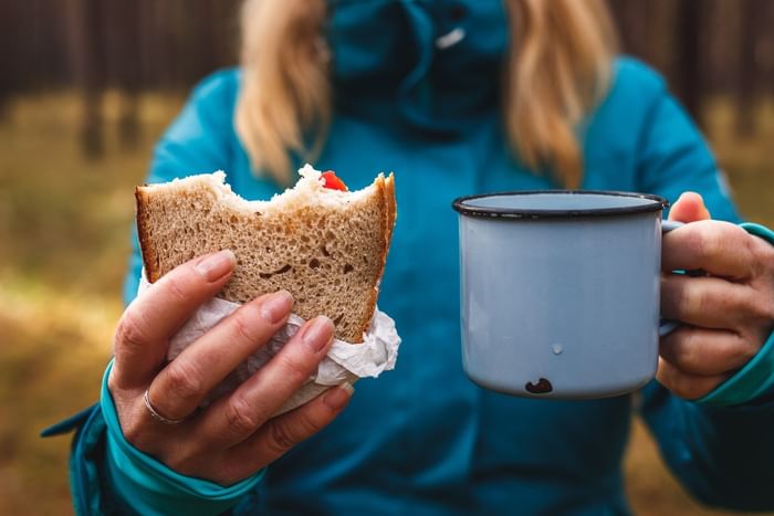 Person in teal jacket holding a sandwich and enamel mug outdoors on a walk in Eastbourne