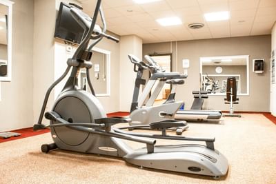 Stationary exercise bikes sit by a treadmill under bright square ceiling lights in a gym at the Rodd Charlottetown Hotel