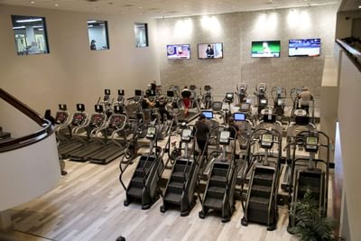 Cardio gym with rows of treadmills and stair climbers beneath four TVs in Miami Lakes Athletic Club at Miami Lakes Hotel