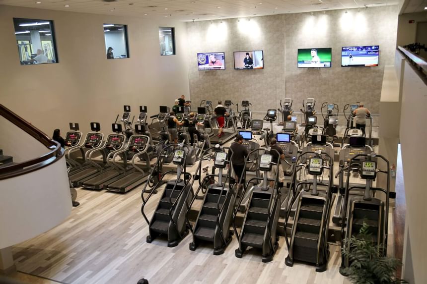 Cardio gym with rows of treadmills and stair climbers beneath four TVs in Miami Lakes Athletic Club at Miami Lakes Hotel