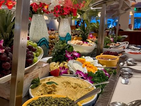 Buffet table with fruits, nuts, hummus, crackers, and flowers, under a clear cover.