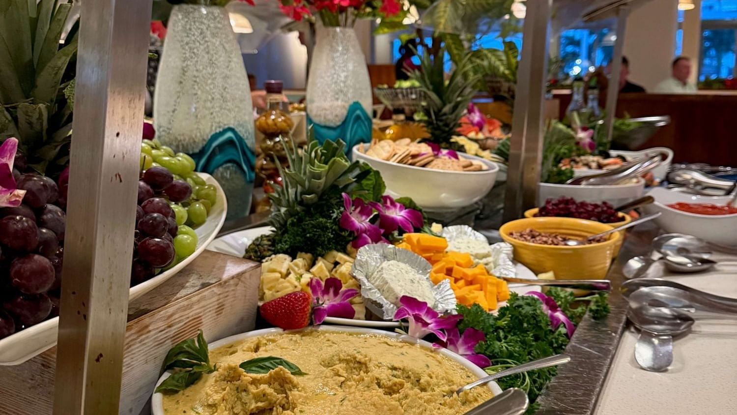 Buffet table with fruits, flowers, hummus, and various salads with spoons on a metal tray