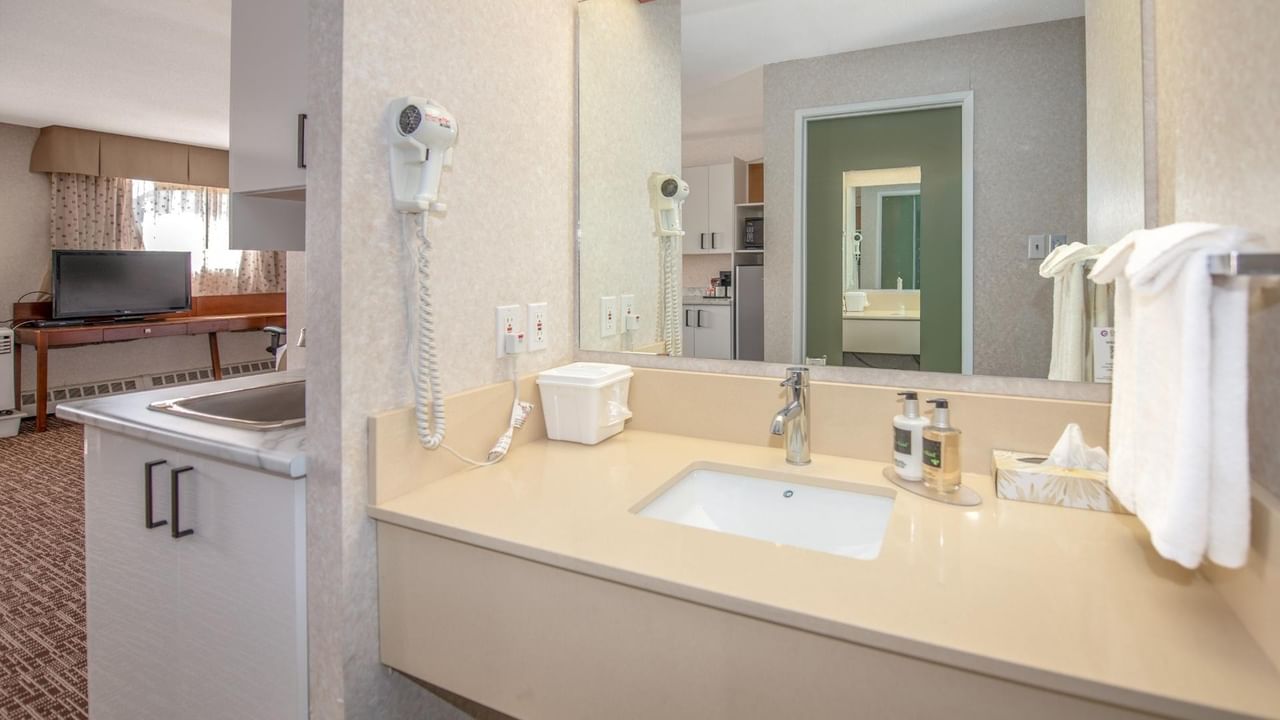 A bathroom with a sink, mirror, hair dryer, towels, and a television in the background.