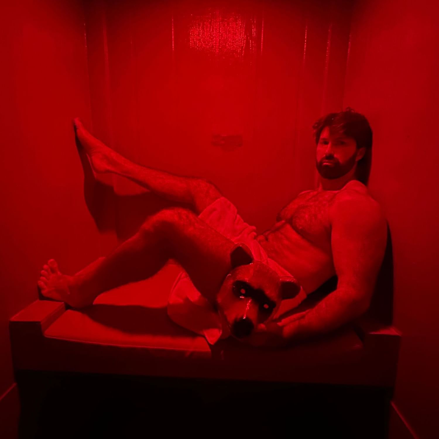 A muscular man reclines in a small, dimly lit sauna room bathed in red light, wearing a towel at The Gaythering