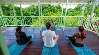 Four guests meditating on blue mats on an elevated wooden balcony overlooking trees at Sugar Cane Club Hotel & Spa