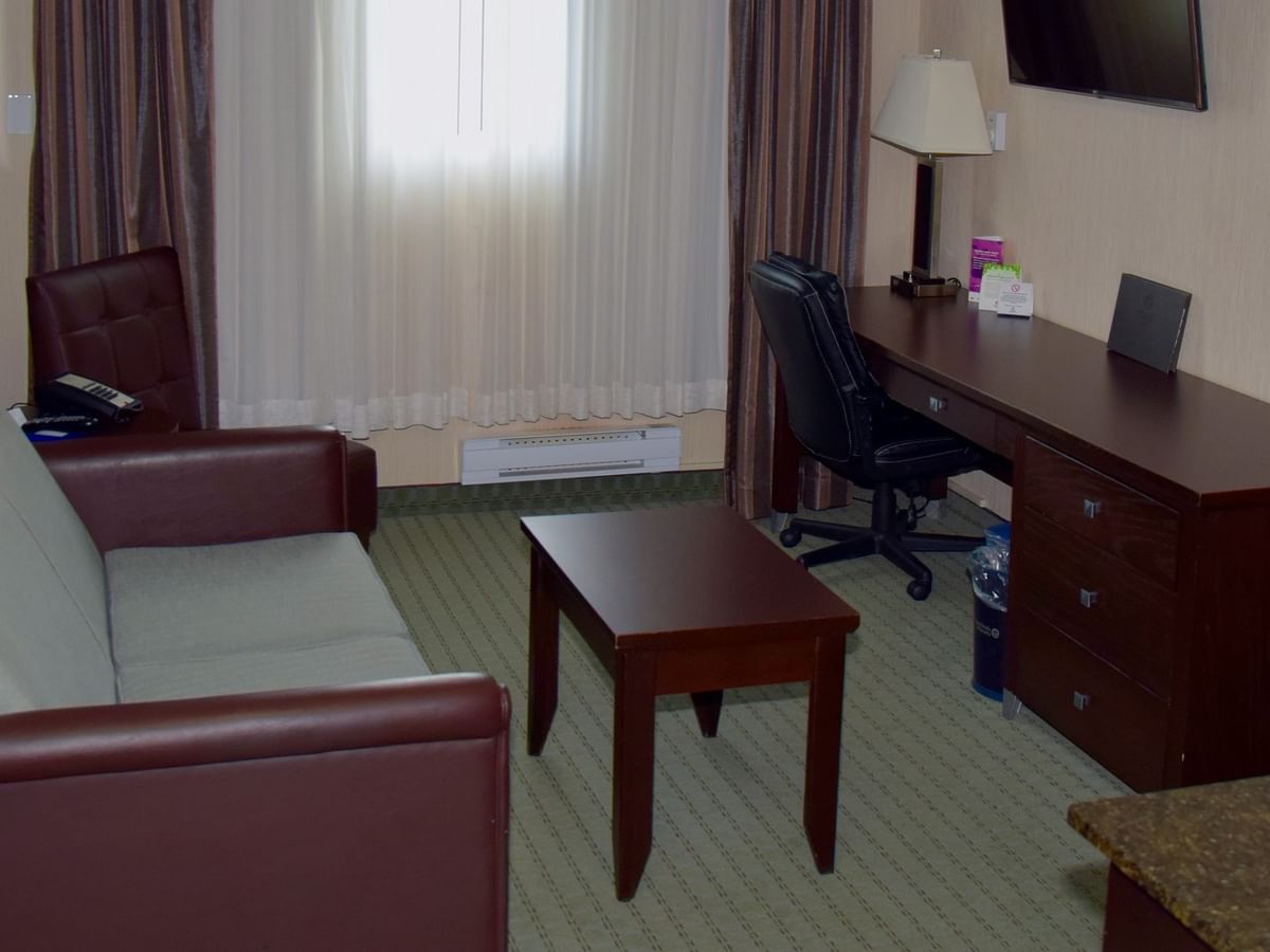 A hotel room with a couch, desk, chair, TV, lamp, and window with curtains.