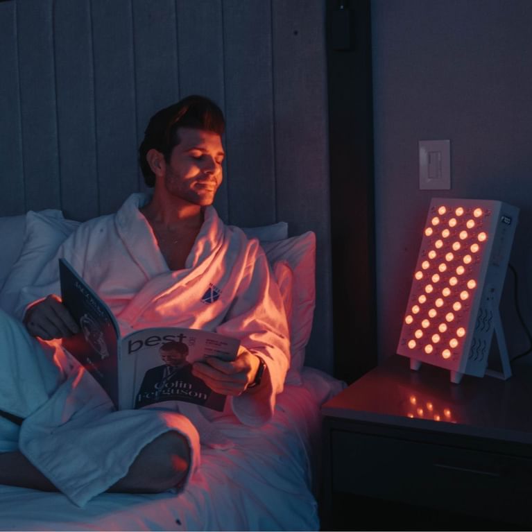Guest in white robe reading a magazine on bed with in-room red light therapy panel