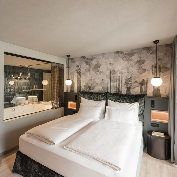 Elegant hotel room with large bed, white linens, decorative pillows, and a bathroom with a large mirror and bathtub.
