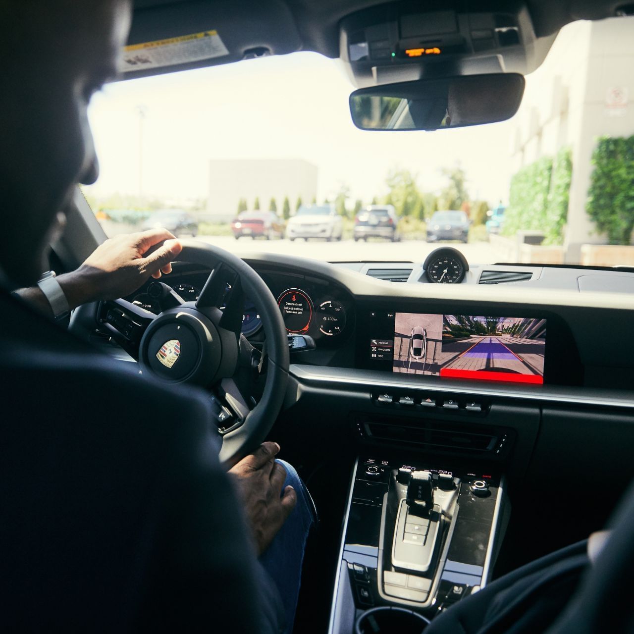 Man driving a car with a rear camera display showing a driveway on the dashboard.