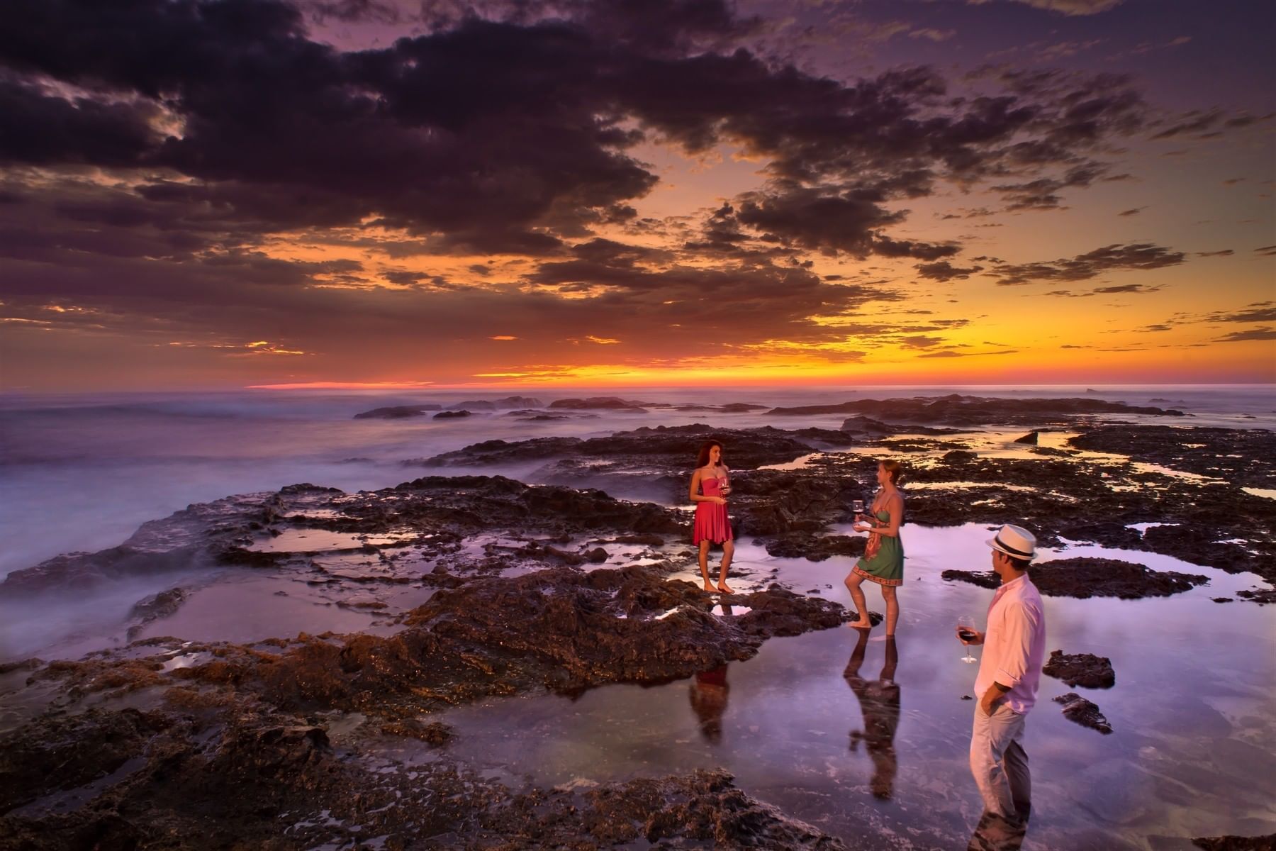Guests enjoy drinks at sunset on the rocky tide pools under a dramatic purple sky near Cala Luna Boutique Hotel