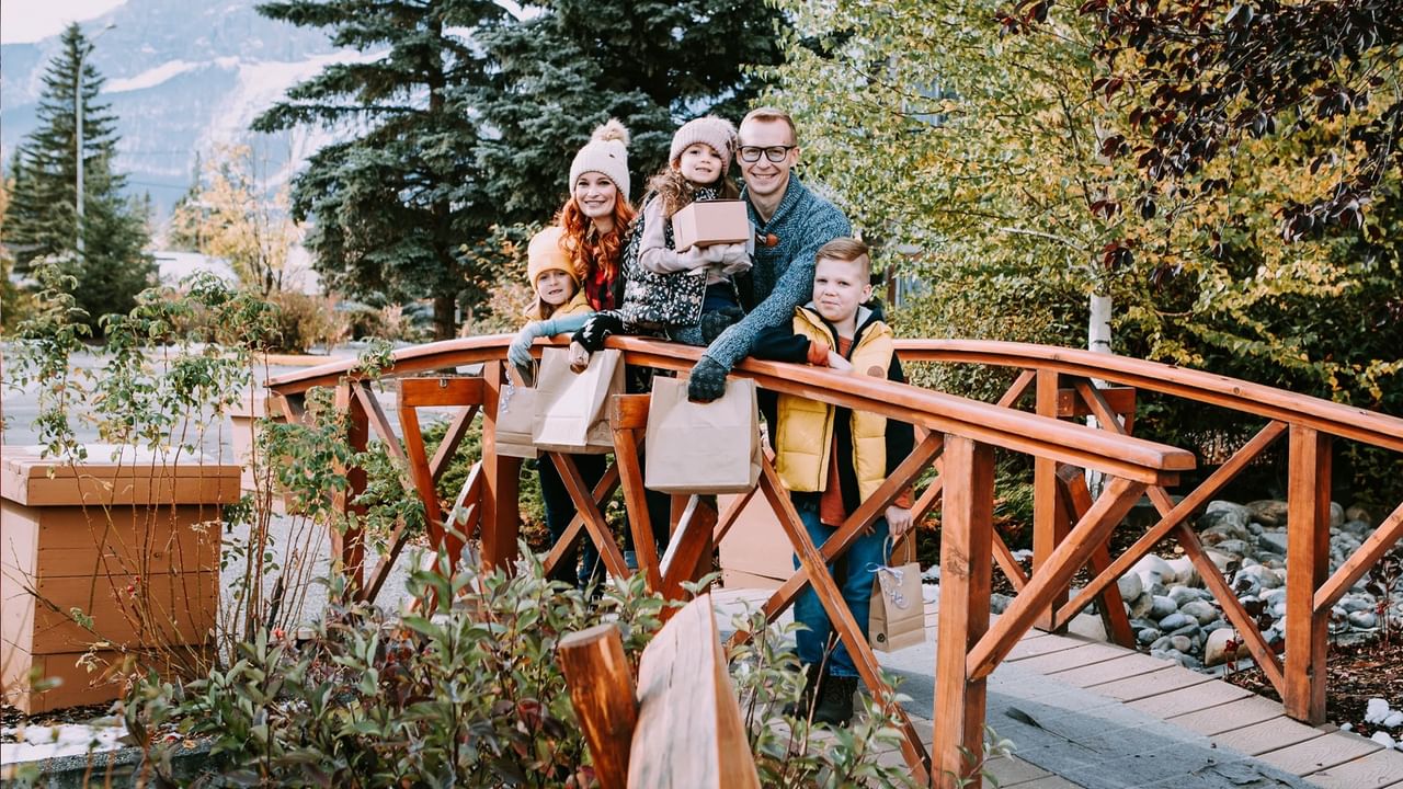 Family of four stands on wooden bridge holding shopping bags with mountains in the background.