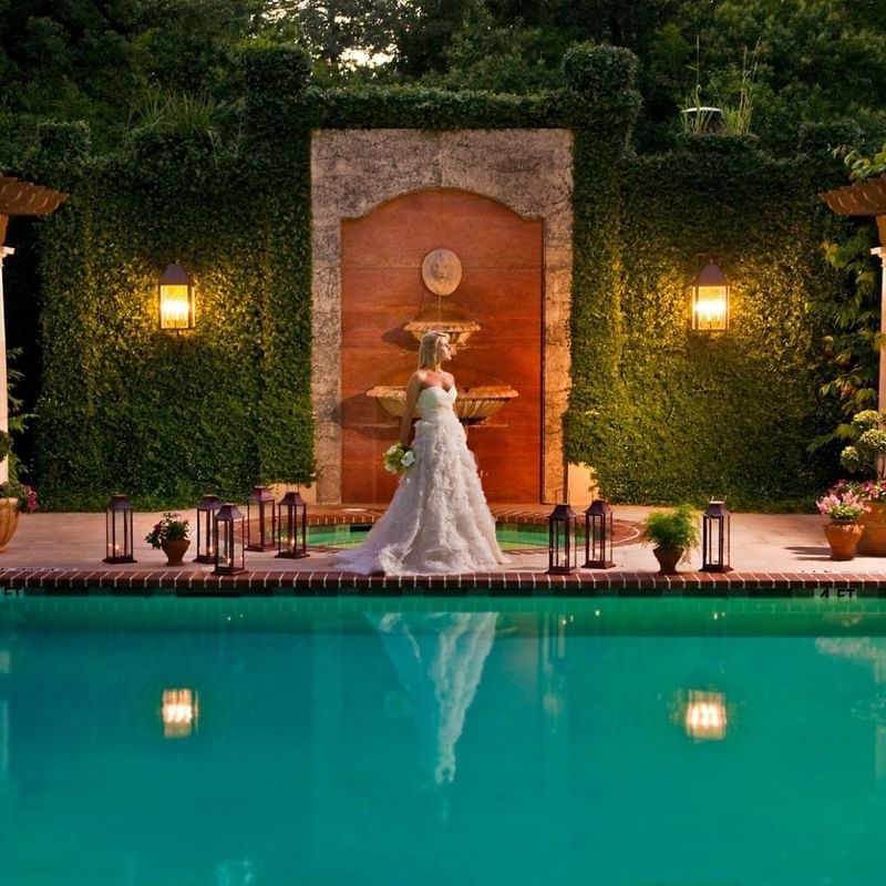 Stunning bride in a flowing white gown stands by a lit pool with a fountain backdrop at Granduca Houston