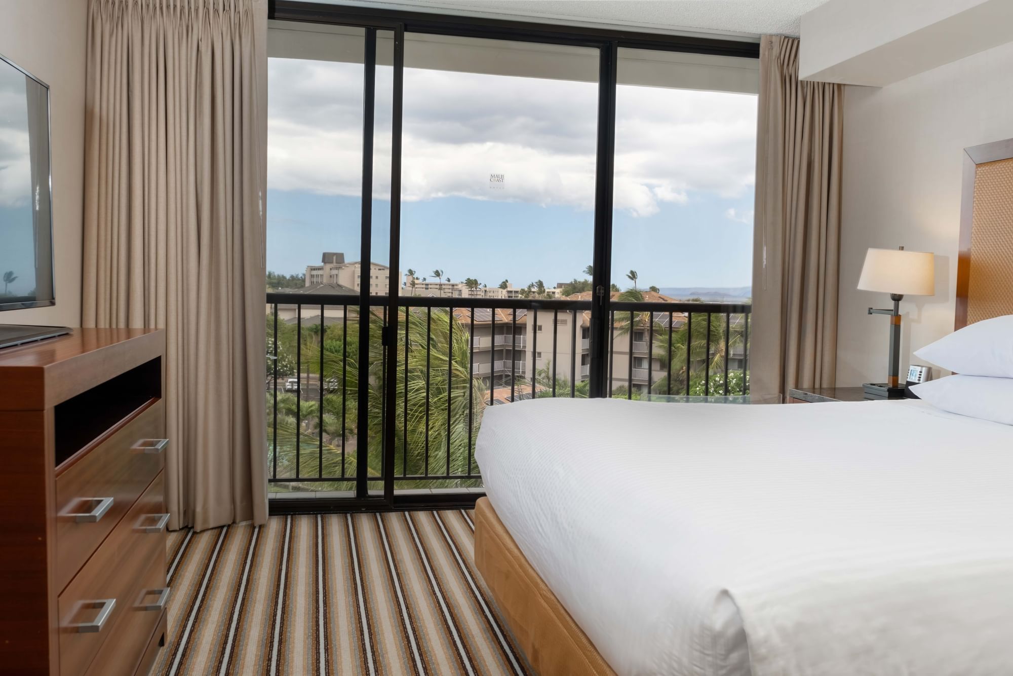 A hotel room with a large bed, TV, and sliding glass doors leading to a balcony with a view.