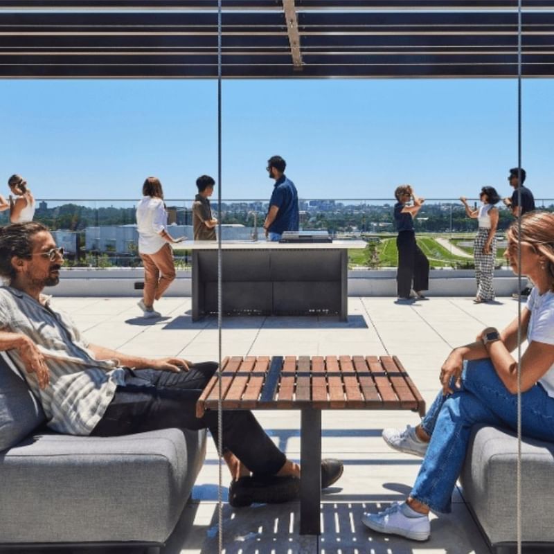 Two people sit on couches by a glass wall, with others standing outside on a rooftop.