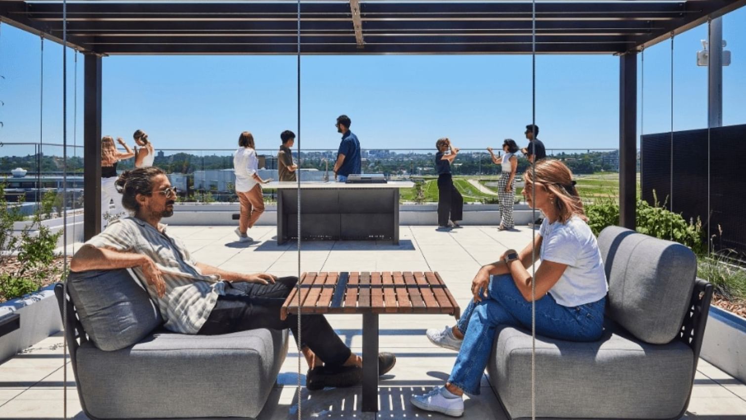 Two people sit on couches by a glass wall, with others standing outside on a rooftop.