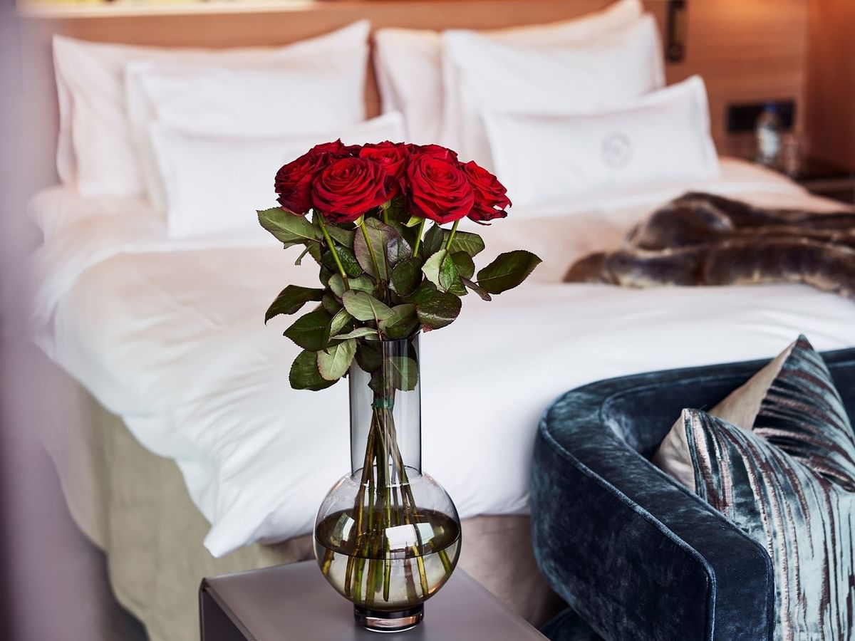 En vase med røde roser på et bord ved siden af en seng og en stol på et hotelværelse.