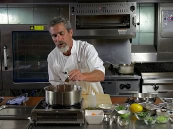 Chef in uniform adding ingredients to a metal pot on an induction cooktop in a commercial kitchen setup at Margaux Restaurant