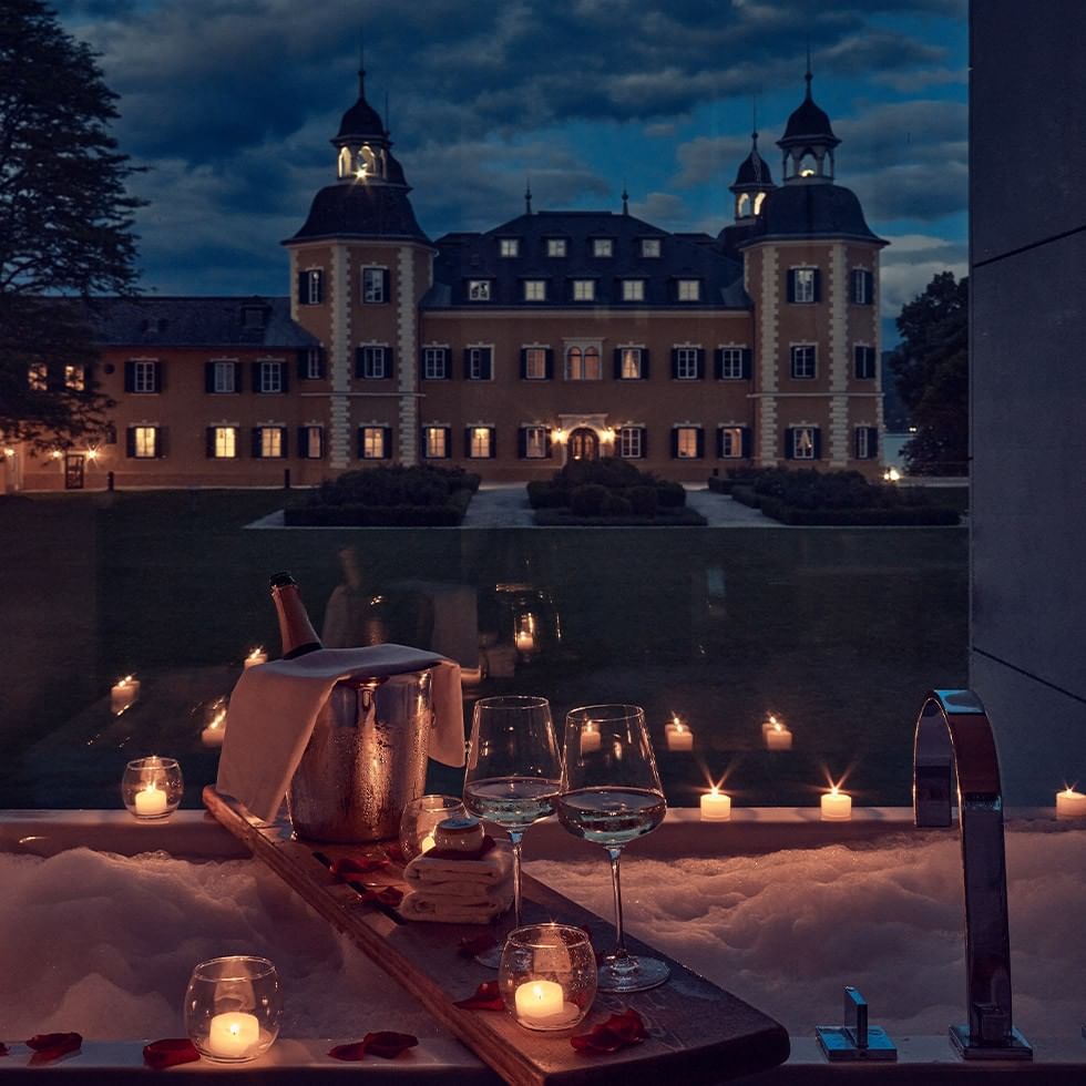 A luxurious hotel room with a large bathtub filled with candles and wine glasses, overlooking a grand building.