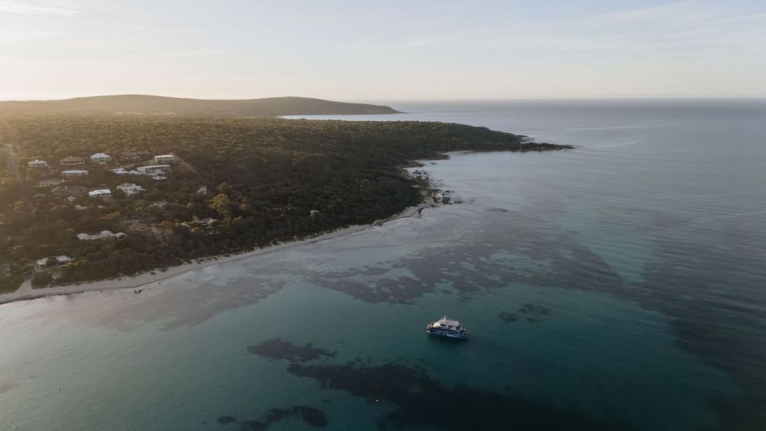 Aerial view of a white boat anchored in turquoise coastal waters near a forested shoreline at golden hour in Margaret River