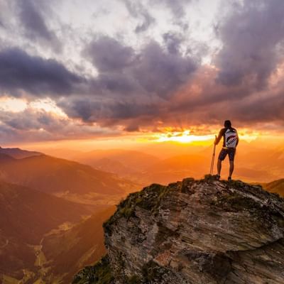 Man in a white and red t-shirt stands on a mountain peak at sunset.