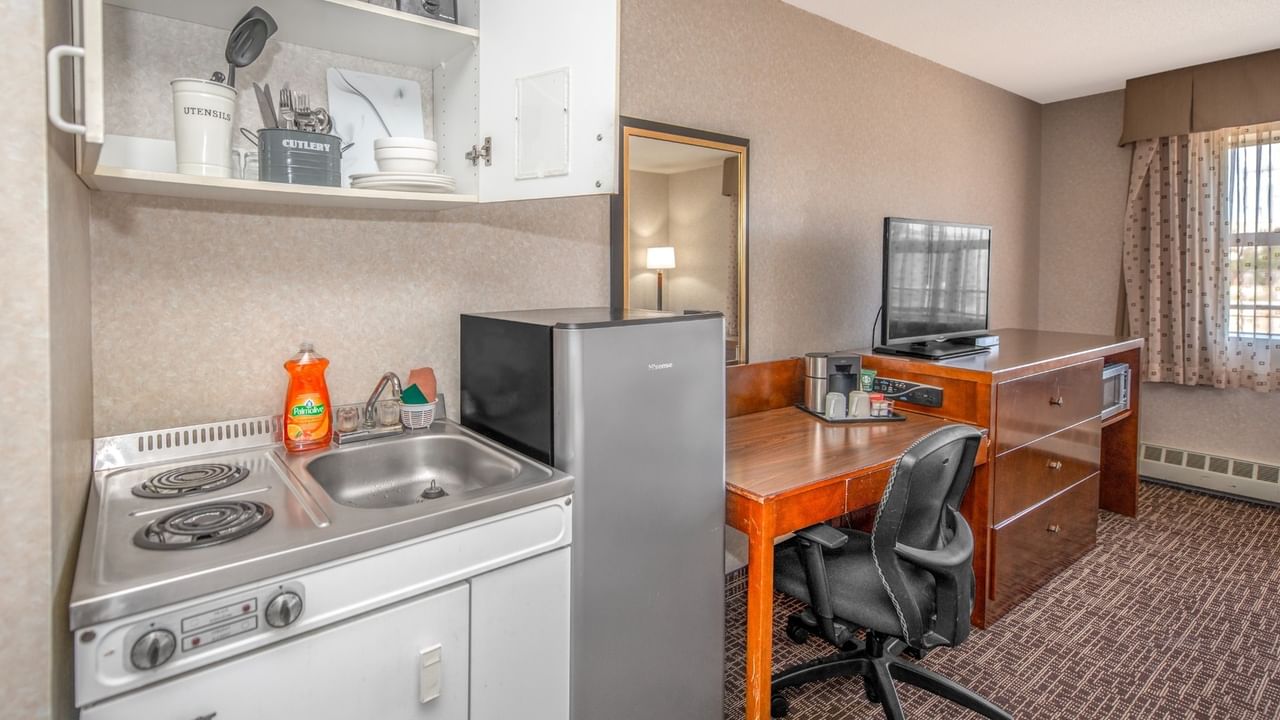 A room with a kitchenette, refrigerator, desk with chair, and a television on top of a dresser.